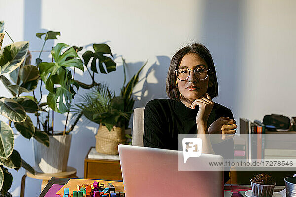 Geschäftsfrau mit Hand am Kinn  die zu Hause am Laptop sitzt