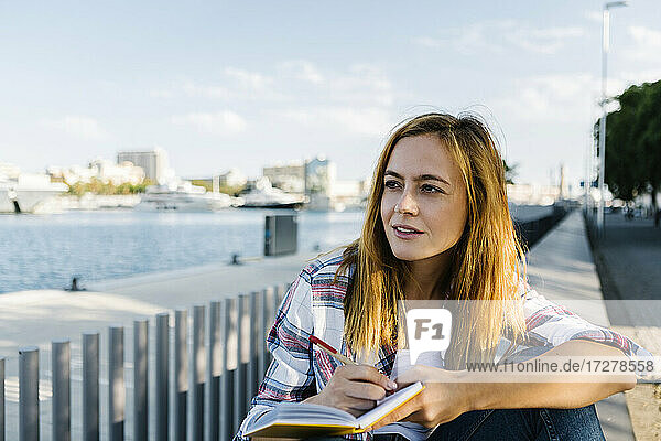 Nachdenkliche junge Frau mit Buch auf dem Fußweg am Meer sitzend