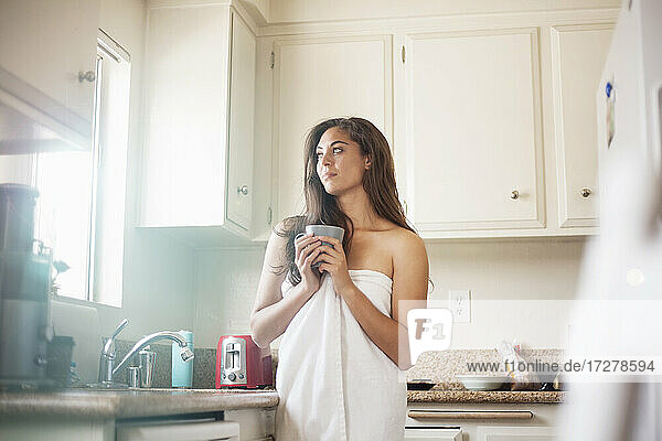 Thoughtful woman wearing towel holding coffee cup while standing in kitchen at home