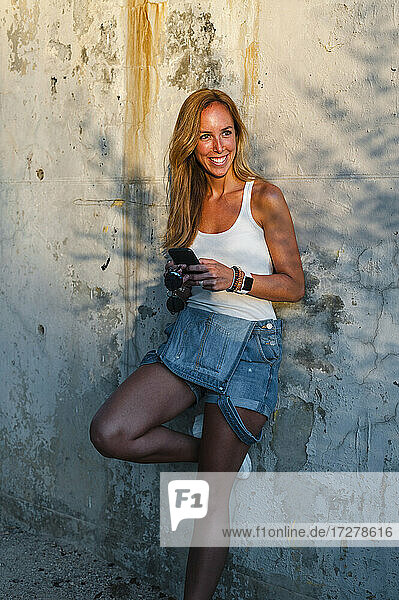 Lächelnde erwachsene Frau  die ein Mobiltelefon benutzt und sich an eine Wand lehnt