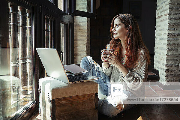 Junge Frau trinkt Kaffee  während sie zu Hause am Laptop arbeitet