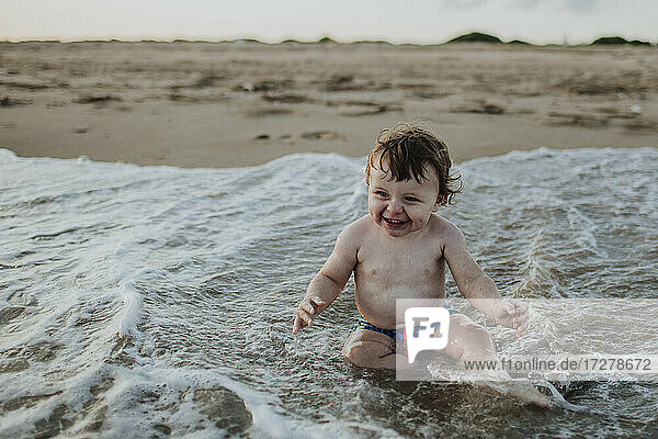 Fröhlicher kleiner Junge spielt im Wasser am Strand bei Sonnenuntergang
