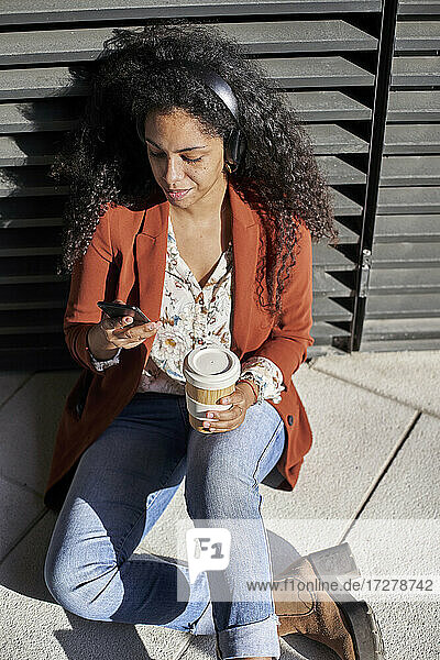 Junge Frau  die ein Smartphone benutzt und eine Kaffeetasse hält  während sie auf einem Fußweg sitzt