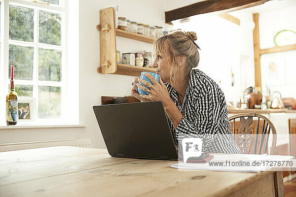 Smiling senior woman looking away while drinking coffee sitting with laptop in kitchen at home