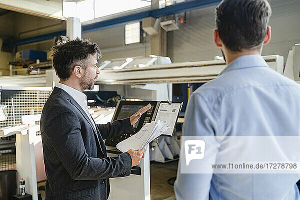 Geschäftsmann mit Papier im Gespräch mit einem Kollegen  während er in einer Fabrik an einer Maschine steht