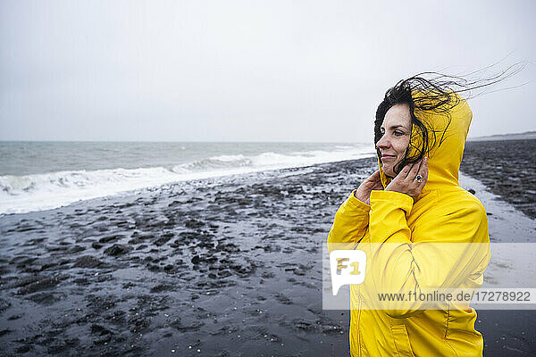 Smiling mature woman in yellow raincoat looking at view while standing on sea shore