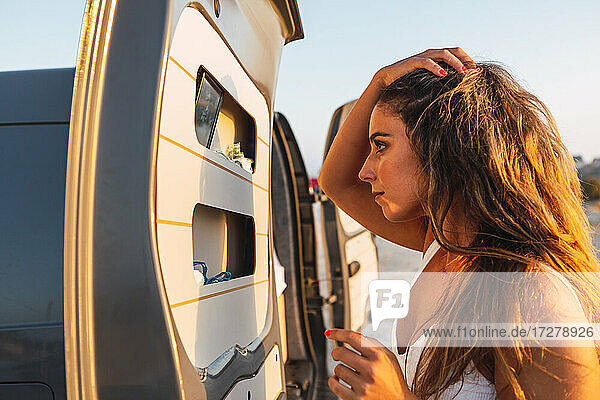 Young woman with hand in hair looking at mirror in camper van door while standing at beach