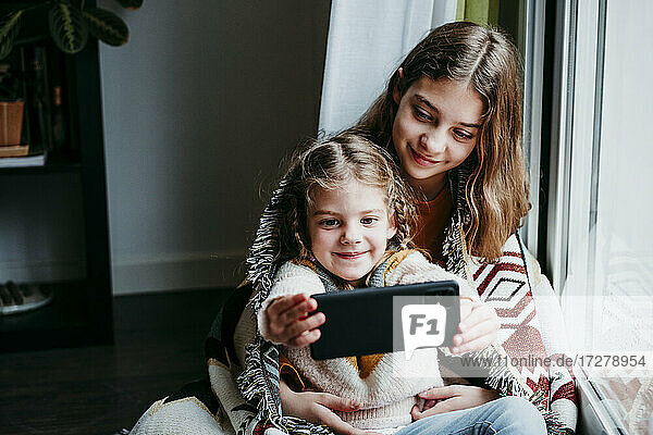 Girl taking selfie with sister while sitting by window at home