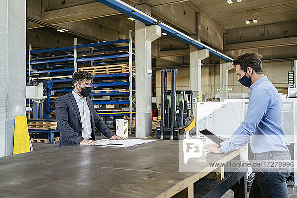 Businessman wearing face mask using digital tablet while standing at social distance with colleague in factory