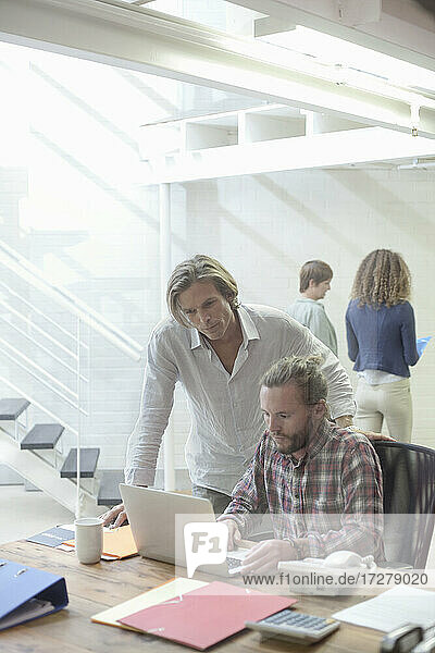 Mann arbeitet an einem Laptop mit einer Mitarbeiterin im Hintergrund im Büro