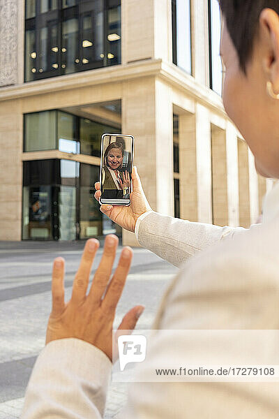 Woman waving hand to friend on video call through mobile phone while standing in city