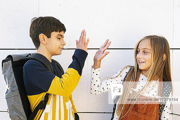 Bruder und Schwester tun hohe fünf  während stehend gegen weiße Wand auf sonnigen Tag