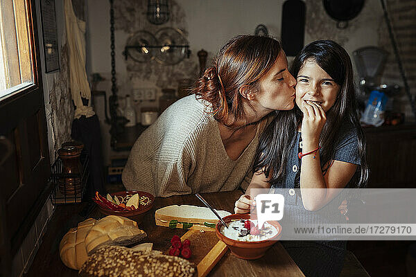 Mother kissing daughter while standing in kitchen at home