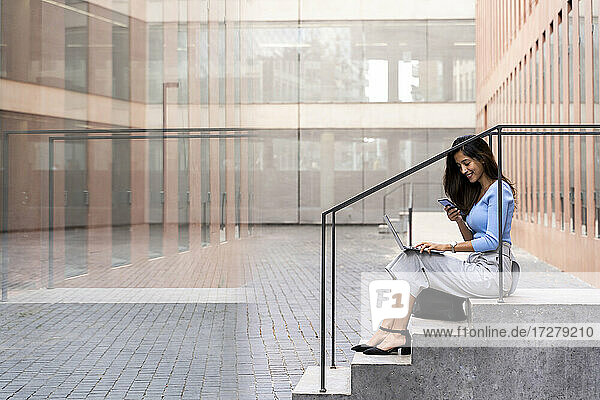 Lächelnde Geschäftsfrau  die ein Smartphone benutzt  während sie mit einem Laptop auf den Stufen vor einem Bürogebäude sitzt