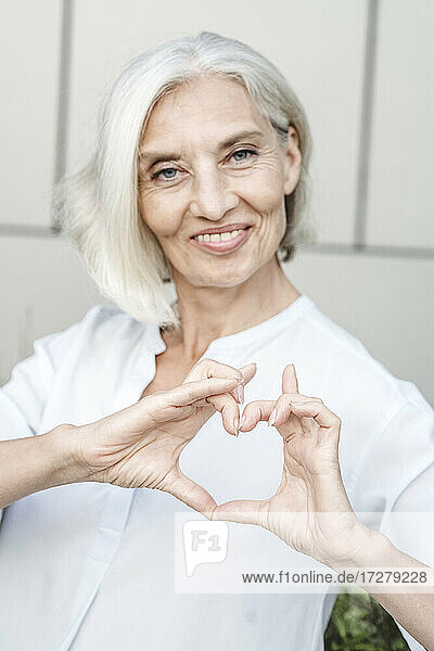 Smiling businesswoman making heart shape with finger while standing against wall