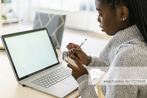 Junge Frau schreibt in einen Notizblock  während sie zu Hause am Laptop lernt