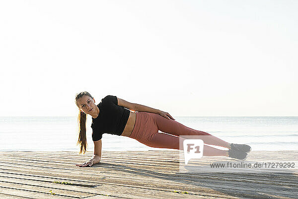 Junge Sportlerin macht Seite Planke Pose auf Promenade gegen klaren Himmel