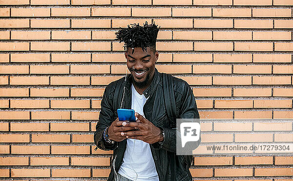 Lächelnder mittelgroßer erwachsener Mann in Lederjacke  der ein Mobiltelefon benutzt  während er an einer Backsteinmauer steht