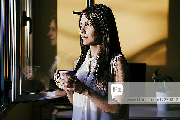 Mittlere erwachsene Frau schaut durch das Fenster  während sie mit einer Kaffeetasse zu Hause steht