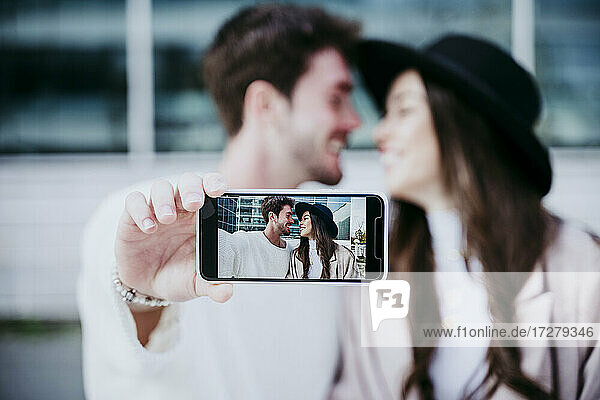 Lächelnder Mann  der ein Foto mit seinem Smartphone macht  während er seine Partnerin in der Stadt betrachtet