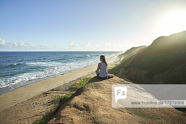 Weibliche Touristin sitzt auf einer Felsformation und schaut aufs Meer