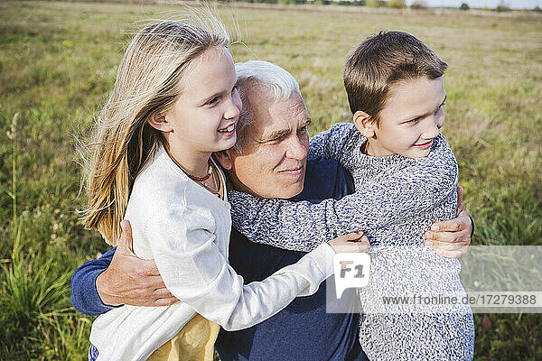 Grandfather looking away while embracing cute grandchildren on field