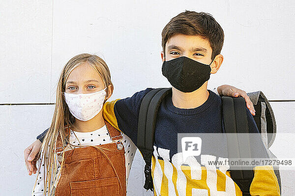 Bruder und Schwester mit Schutzmaske an der Wand stehend an einem sonnigen Tag
