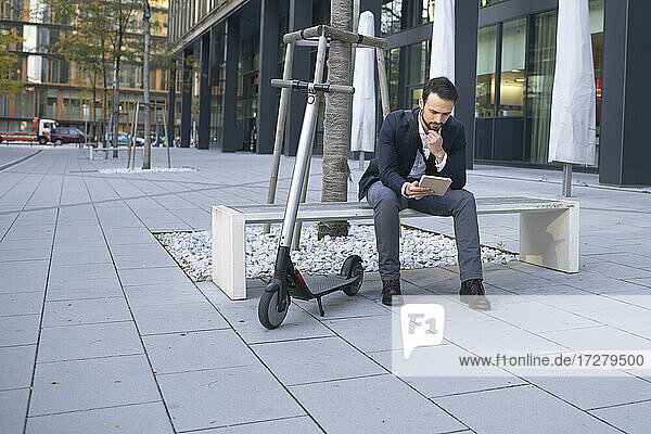 Geschäftsmann  der ein digitales Tablet benutzt  während er mit einem E-Scooter auf einer Bank in der Stadt sitzt