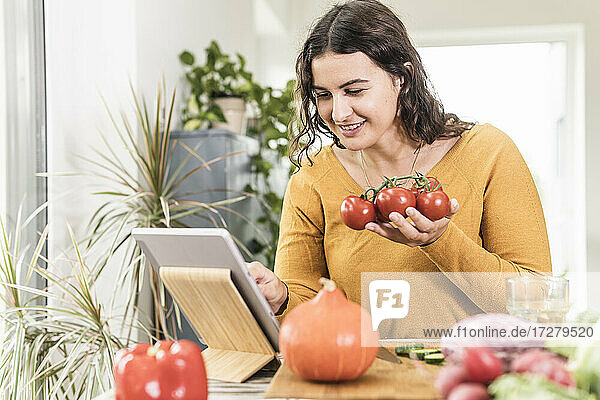 Lächelnde Frau  die eine Tomate in der Hand hält und ein digitales Tablet zu Hause benutzt