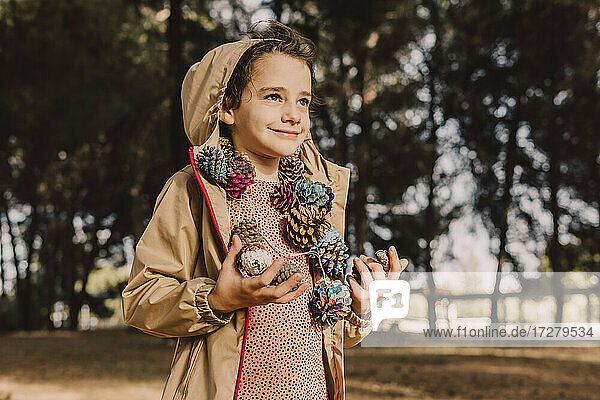 Lächelndes niedliches Mädchen mit bunter Tannenzapfengirlande schaut weg  während sie im Park steht