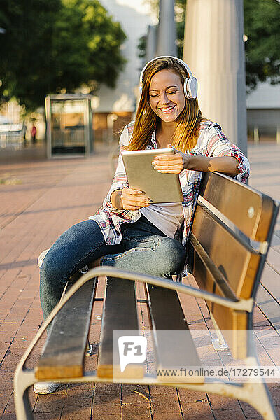 Lächelnde Frau  die ein digitales Tablet benutzt  während sie an einem sonnigen Tag auf einer Bank sitzt