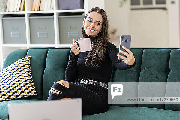 Lächelnde schöne weibliche Bloggerin  die ein Selfie mit Kaffeetasse macht  während sie auf dem Sofa zu Hause sitzt