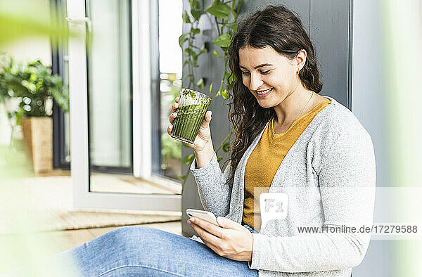 Junge Frau  die ein Mobiltelefon benutzt  während sie zu Hause einen Smoothie trinkt