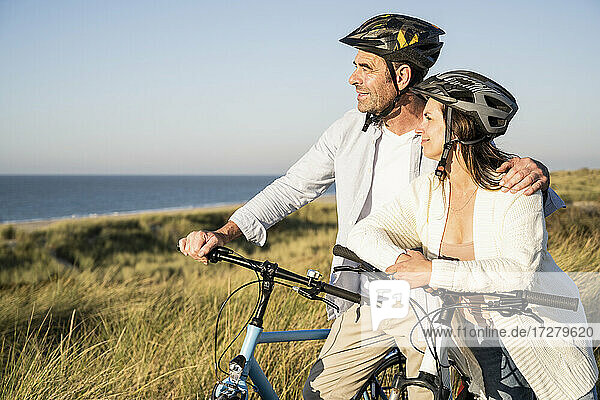 Lächelndes reifes Paar  das die Aussicht betrachtet  während es mit Fahrrädern am Strand steht
