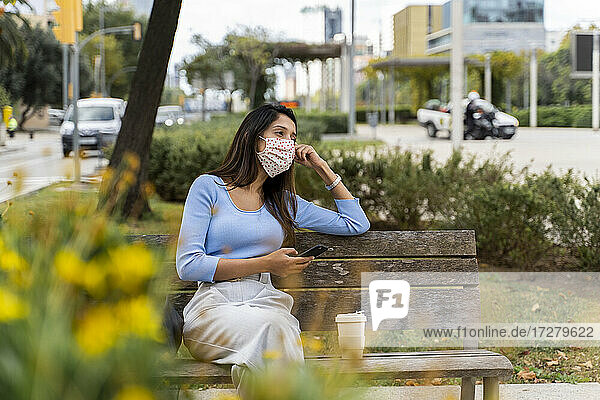Nachdenkliche Frau  die mit ihrem Smartphone wegschaut  während sie während COVID-19 auf einer Bank in der Stadt sitzt
