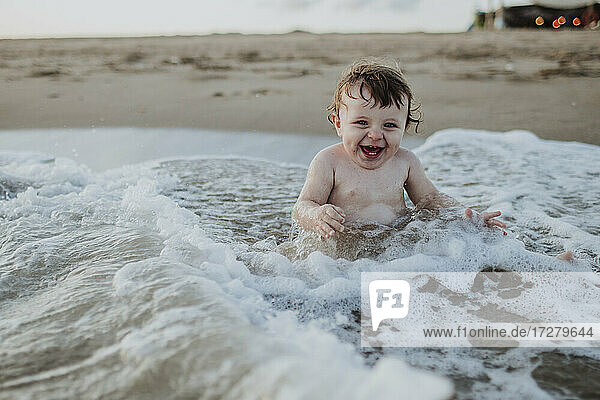 Männliches Kleinkind lachend im Wasser sitzend am Strand bei Sonnenuntergang