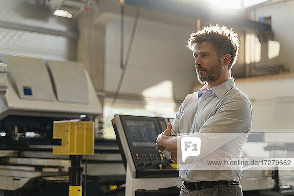 Mature businessman standing with arms crossed by machine at factory