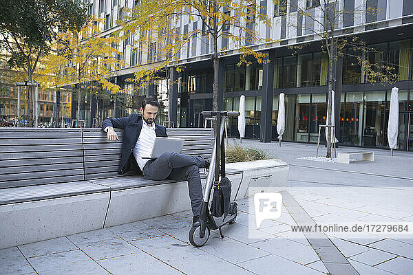 Businessman looking at laptop while sitting by push scooter in city