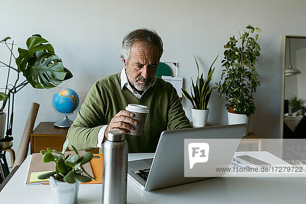 Älterer Mann trinkt Kaffee  während er zu Hause am Laptop arbeitet