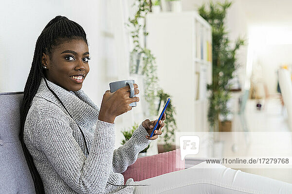 Young woman drinking coffee while using smart phone sitting on sofa in living room at home