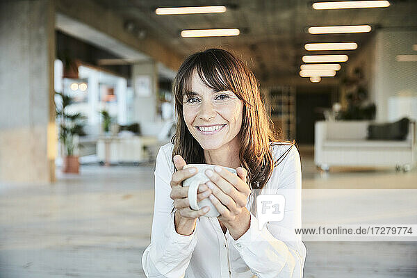 Lächelnde reife Frau hält Kaffeetasse  während sie zu Hause sitzt