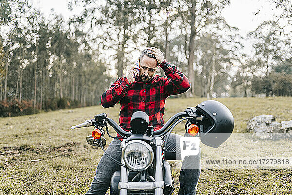 Angespannter männlicher Motorradfahrer,  der auf einem Motorrad im Wald sitzt und mit seinem Smartphone telefoniert
