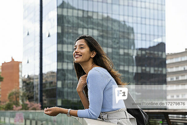 Lächelnde Geschäftsfrau  die wegschaut  während sie sich an ein Geländer eines modernen Gebäudes lehnt