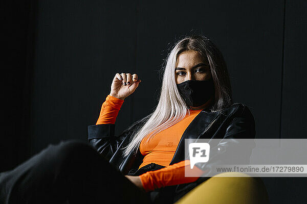 Junge Frau mit Gesichtsschutzmaske sitzt auf einem Sofa vor einer schwarzen Wand