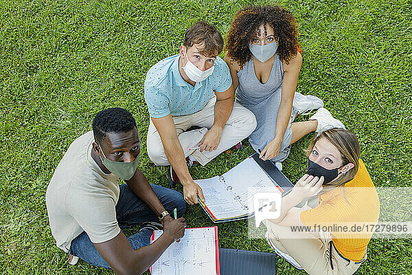 Studenten mit Schutzmaske  die auf dem Rasen eines Universitätsgeländes zusammensitzen und lernen