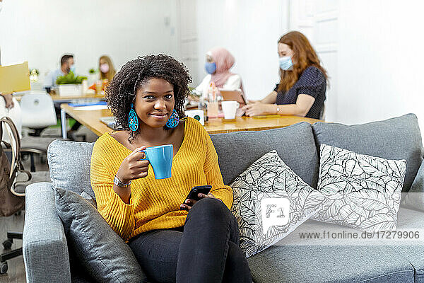 Smiling woman drinking coffee while sitting with coworker working in background at office