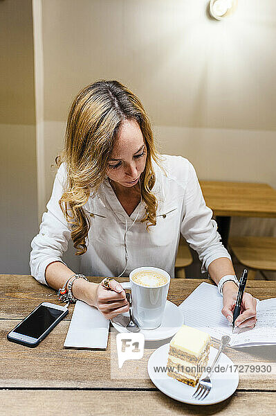 Geschäftsfrau schreibt in ein Notizbuch  während sie Kaffee und Karottenkuchen am Tisch im Büro genießt