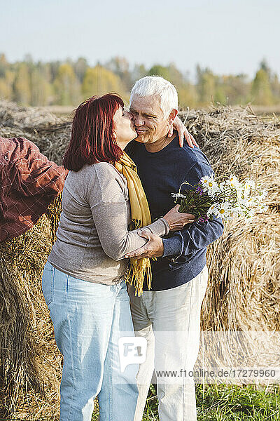 Glückliches älteres Paar  das sich küsst  während es gegen Heuballen auf einem Feld steht