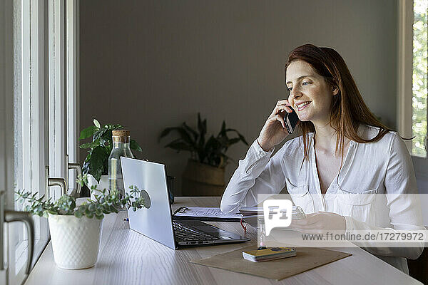 Lächelnder Arzt  der mit seinem Handy telefoniert  während er zu Hause am Laptop arbeitet