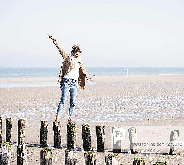 Lächelnde junge Frau mit ausgestreckten Armen  die an einem sonnigen Tag auf Holzpfählen am Strand spazieren geht